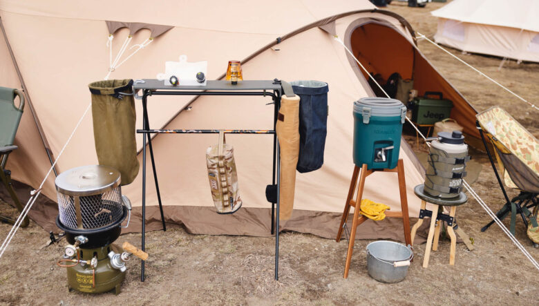 キャンプの定番となりつつあるミニマルワークスのインディアンハンガーに、キャンプギークスの天板を取り付け、機能を拡張。天板左に取り付けた金具はダルトンのもので、輪っか部分に袋を取り付けゴミ箱として使える。