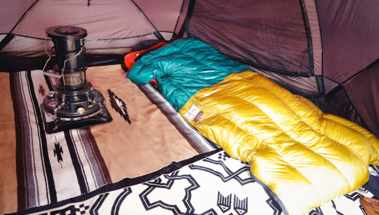 テント室内は、ラグを敷きアラジンのストーブをインストール。寝袋はナンガ×ネイタルデザインのモデルでカラーリングが特徴的だ。しっかり防寒対策することで広い室内も暖かく。