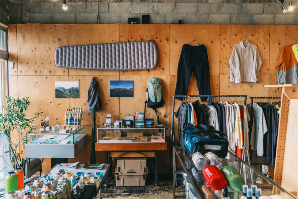 沖縄・宜野湾のアウトドアショップ「narrative outdoor supply」



下書き保存

公開

31/100

82
/100



ブロックタイプまたはスタイルを変更

画像ブロックを37から36へ上方向に移動
画像ブロックを37から38へ下方向に移動
配置を変更


【時松執筆中】海だけじゃない、沖縄の外遊びの新しい入口。宜野湾「Narrative Outdoor Supply」が作るローカルな提案。
米軍基地と沖縄の海がのぞめる宜野湾の通り沿いに、アウトドアギアやウエアを扱うショップ「Narrative Outdoor Supply（ナラティブアウトドアサプライ）」は店を構えている。　

店内に並ぶのは、キャンプ用品やハイクギア、沖縄の街にも馴染むオリジナルウエアまで。アウトドアショップではあるけれど、いわゆる山道具の店とも、キャンプ道具だけの店とも少し違う。外遊びの道具と、グラフィック、ローカルなカルチャーが混ざった空間だ。

沖縄のアウトドアといえば、真っ先に海を思い浮かべる人も多いはず。けれどナラティブが提案するのは、それだけではない。もっと身近に、もっと自由に外へ出て、自分なりの楽しみ方を見つける。沖縄の外遊びに、新しい入口をつくろうとしている店だ。

▶︎画像1 キャプション案
米軍基地と海をのぞむ宜野湾の通り沿いにある「Narrative Outdoor Supply」。外遊びの道具とローカルカルチャーが混ざるショップだ。

焚き火から始まった、「アクティビティからメディテーションへ」という考え方。
沖縄・宜野湾のアウトドアショップ「narrative outdoor supply」オーナー
代表の眞謝さん。グラフィックやプリントの制作活動も、店のオリジナルアイテムにつながっている。
現在ナラティブを代表として運営するのは、眞謝さん。ショップオーナーとして意外にも「MASH UP INK」というシルクスクリーンプリント工房を営み、「TOZOW MAJA / トゾウ マジャ」名義で個展も開催するグラフィックアーティストの顔を持つ。

アウトドアとの距離が近づいたのは、コロナ禍にキャンプ、とくに焚き火にハマったことがきっかけ。火を見ていると、頭のなかのざわつきが静まり、自分が何に惹かれているのか、自然と見えるようになってくる。

そんなキャンプ熱をSNSで発信していたところ、狩猟などにも親しむ共同創業者の大野さんと意気投合し、ナラティブを立ち上げるに至った。山やトレッキングに足を踏み入れるようになったのも、大野さんに誘われて里山へ入ったことがきっかけだったという。

那覇でもやんばるでもなく、宜野湾にある理由。
沖縄・宜野湾のアウトドアショップ「narrative outdoor supply」
アウトドアショップの立地として考えるなら、人が集まる街中か、フィールドに近い場所が多いだろう。沖縄でいえば那覇や、やんばるのような北部エリアだ。

けれど、ナラティブが店を構えたのは、沖縄県中部の宜野湾だった。

理由のひとつは、宜野湾が「街と自然の中間地点」にあること。那覇方面からも訪れやすく、北部の自然豊かなフィールドにも足を延ばしやすい。眞謝さん自身の活動拠点がもともと宜野湾だったこともあり、ここが現実的で動きやすい場所だったという。

沖縄・宜野湾のアウトドアショップ「narrative outdoor supply」オーナー
そして、この立地は、店が提案したい外遊びともつながっている。最近始めているのが、店から読谷方面まで歩き、ビーチでテント泊をして戻ってくるような「シティハイク」だ。

沖縄は車移動が当たり前で、長い距離を歩くことが文化としてあまり根付いてはおらず、珍しく見える土地でもある。だからこそ、あえて街を歩く。途中で飲食店に立ち寄ったり、写真を撮ったりしながら歩いて、ビーチでテントを張る。観光でも登山でもない、沖縄の新しい遊び方がそこにはある。

▶︎画像3 キャプション案
宜野湾は、街にも自然にもアクセスしやすい中間地点。店の提案する外遊びとも相性がいい。

古いコインランドリーに、アウトドアギアと工房を詰め込んで。
沖縄・宜野湾のアウトドアショップ「narrative outdoor supply」オーナー
古い建物の質感を活かした店内。木の什器が、アウトドアギアの並びに温かさを加えている。
店舗が入る建物は、もともとコインランドリーだった場所。天井は高く、ところどころに古い建物らしい荒さも残っている。

内装で目を引くのは、三角形の什器や壁面に使われた針葉樹合板。眞謝のイメージしたものを、先輩の家具職人にワンオフで作ってもらったものだという。古い建物の無機質さに、木の温かさが加わって、店内には独特の落ち着きがある。

沖縄・宜野湾のアウトドアショップ「narrative outdoor supply」オーナー
店の奥には「MASH UP INK」のプリント工房も併設。オリジナルアイテムを自分たちの手で作れるのも特徴だ。
店舗の奥には、眞謝さんが長年続けてきたシルクスクリーン工房「MASH UP INK」がある。今年からこの場所へ移転し、ナラティブの店内でプリント作業まで行えるようになった。

元店長が独立して始めた刺繍の作業場も同じ空間にあり、キャップやウエアの刺繍、プリントまで、店の中で形にできるのがこの場所の強みだ。セレクトショップでありながら、自分たちで作ることもできる。ナラティブの空間には、売る場所と作る場所が地続きになっている面白さがある。

外遊びを、メディテーションの時間へ。
沖縄・宜野湾のアウトドアショップ「narrative outdoor supply」オーナー
店名の「Narrative」には、自分だけの体験や物語を育てていくような意味が込められている。

眞謝さんが掲げるのは、「アクティビティからメディテーションへ」という考え方だ。ここでいうメディテーションとは、特別な作法としての瞑想ではない。焚き火を眺めたり、山を歩いたり、目の前の自然や身体の動きに意識を向けることで、頭のなかにある余計な思考を少しずつほどいていく。そんな時間を指している。

﻿

自分の感覚がクリアになると、何に惹かれるのか、自分にとって何が心地いいのかも見えやすくなる。外で遊ぶことを通して、自分なりの基準を知る。その先で、道具の選び方や日々の過ごし方まで少しずつ変わっていく。

ナラティブが提案しているのは、ただアクティブに動くための外遊びではない。自然のなかで過ごす時間を、自分自身と向き合うきっかけにもしてほしい。そんな思いが、店名にも、店頭に並ぶ道具にも通っている。

道具はシンプルに。目の前の自然を楽しめるセレクト。
画像
画像ファイルをアップロードするか、メディアライブラリから選択、または URL を追加してください。
アップロード
メディアライブラリ
URL から挿入
ナラティブのセレクトで眞謝さんが意識しているのは、シンプルで安心感があり、初めて使う人にも扱いやすいものだという。

それは、目の前の自然を楽しむために、道具の扱いで頭のキャパを使いすぎてほしくないから。使い方が複雑すぎず、フィールドで素直に頼れるもの。Rab（ラブ）やEXPED（エクスペド）など、ベーシックな海外ブランドのアイテムが多いのも、その考え方とつながっている。

﻿

一方で、沖縄ならではの気候や暮らし方もセレクトには表れている。近年は本土でも、アウトドアブランドのシェルが山道具としてだけでなく、街で着るファッションアイテムとして浸透してきた。

車移動が多く、雨が降っても傘を差さない人が多い沖縄なら、雨をしのげて見た目もいいウインドシェルやレインウエアは、もっと日常に馴染むはず。眞謝さんは、そうしたアイテムが沖縄の街でも自然に選ばれるようになる可能性を感じているという。

▶︎画像6 キャプション案
RabやEXPEDなど、扱いやすさと信頼感のあるブランドも揃う。初めての道具選びにも入りやすい。

オリジナルもセレクトも、街と外遊びを行き来するものを。
﻿

ナラティブの面白さは、セレクトだけでなく、自分たちで作れることにもある。店の奥にはプリント工房があり、刺繍の作業場も隣接。オリジナルウエアやキャップには、グラフィックやディテールを自分たちの手で形にできる店ならではの強みが出ている。

店頭でもまず目を引いたのは、オリジナルのセットアップやTシャツ類。一方で、店頭には外へ出る気分をアゲてくれる小物も並んでいた。

沖縄の街とフィールドを行き来する、オリジナルの2WAYセットアップ。
﻿

オリジナルアイテムの中でも存在感があったのが、「Narrative Original / 2Way Shirts」と「Narrative Original / 2Way Guide Pants」。どちらも透湿性・簡易防水を備えたナイロン100％の生地を使い、街でも外遊びでも着やすいバランスに仕上げたウエアだ。

﻿

シャツは、袖をファスナーで取り外せる2WAY仕様。長袖としても半袖としても着られるので、日差しの強い時期や、旅先での温度変化にも対応しやすい。フロントポケットをあえて排除し、背面には大容量のゲームポケットを配置。ザックを背負ったときの干渉を避けつつ、街歩きや移動中でも手ぶらで動きやすい作りになっている。

﻿

パンツも同じく2WAY仕様で、左右の裾を取り外せばハーフパンツとして着用可能。ジップ付きポケットや裾のドローコードなど、移動中や外遊びで使いやすいディテールも備える。

セットアップで着れば、街、旅、フィールドをそのまま行き来できる。沖縄の気候や街の空気にも馴染みつつ、外で使うことまで考えられた、ナラティブらしいウエアだ。

▶︎画像7 キャプション案
袖や裾を取り外せる、Narrative Outdoor Supplyの2WAYセットアップ。街にも外遊びにも馴染む一着。

▶︎画像8 キャプション案
ボディから作り込んだNarrative Outdoor Supplyのオリジナルキャップ。長めのツバと刺繍が印象的で、街歩きにも外遊びにも取り入れやすい。

リフレクターのグラフィックが効いた、Parallel Teeシリーズ。
﻿

ナラティブのオリジナルTシャツ「Parallel」シリーズもナラティブのアイコン的アイテム。グラフィックを主役にしながら、夜道やアクティビティでの視認性にも配慮されているのが特徴だ。

長袖の「Parallel L/S T-shirts」は、光が当たると反射するリフレクティブプリントを採用。吸汗速乾性やUVカット機能を備えたポリエステル100％の生地で、タウンユースはもちろん、ラッシュガード的にも使いやすい。

﻿

半袖の「Parallel Tee 4th」は、4周年記念で作られたモデル。チャコールのボディにリフレクターインクを使ったグラフィックが入り、車のライトやカメラのフラッシュに反応する。プリント工房を持つナラティブらしさが出た一枚だ。

﻿

なお、現在はオリジナルのバンダナスカーフも再生産中だという。沖縄のヴィンテージマップをベースに、タトゥーアーティストのKENTA氏がデザインを手がけた大判スカーフで、再入荷したらこちらもチェックしたい。

▶︎画像9 キャプション案
リフレクティブプリントを取り入れた「Parallel」シリーズ。グラフィックと機能を同じ温度で楽しめるTシャツだ。

▶︎画像10 キャプション案
再生産中のオリジナルバンダナスカーフ。ヴィンテージの沖縄地図をベースにした、スーベニア感のある一枚。

ネオンカラーのパラコードが映える、PIQNICのボトルホルダー。
﻿

セレクトアイテムでは、PIQNIC（ピクニック）の「Bottle Holder Bag 001」も印象的だった。パラコードを編み込んだような立体感と、ネオンカラーの配色が目を引く、ハンドメイドのボトルホルダーだ。

カラビナ付きでギア感もあり、街歩きやフェス、ビーチへの外出にも持ち出しやすい。500mlのペットボトルから1.0Lのナルゲンボトルまで対応し、ストラップの長さやショルダーカバーも調整できる。

﻿

水分補給の道具をバッグの中にしまい込まず、すぐ手に取れる場所に置いておけるのが便利。実用的でありながら、アクセサリー感覚でも楽しめる。こういう小物が自然に並んでいるところにも、ナラティブのセレクトの幅が出ている。

キャプション案
ネオンカラーのパラコードとカラビナが目を引く、PIQNICのボトルホルダー。500mlペットボトルから1.0Lナルゲンまで対応。

ファミリーキャンプから、沖縄の外遊びを広げる店へ。
﻿

オープン当初のナラティブは、ファミリーキャンプ向けのアイテムも多く扱っていたという。キャンプブームの空気もあり、沖縄で外遊びに興味を持つ人の入口として、間口を広げる意味もあったのだろう。

﻿

ただ、現在は少しずつ方向性を見直しているそうだ。より軽く、コンパクトで、街歩きや低山トレッキング、旅先での外遊びにも馴染む道具へ。眞謝さんが今考えているのは、沖縄でアウトドアを楽しむ選択肢を、海やキャンプ以外にも広げていくような売り場づくりだ。

﻿

それは、単に山岳系に振り切るということではない。沖縄で歩くこと、低山を楽しむこと、街から海へ向かうこと。車移動が当たり前の土地で、あえて自分の足で移動してみること。そうした体験に合う道具を、これからもっと増やしていきたいという。

ナラティブは、いままさに沖縄のアウトドアを別の角度から見せようとしている途中にある。

小さなコミュニティが生まれるイベントを増やしていく。
﻿

今後について聞くと、眞謝さんは「小さなコミュニティができるようなイベントを増やしていきたい」と話してくれた。

これまでにも、ビーチで音楽を流しながらキャンプを楽しむイベントなどを行ってきたという。けれど今は、もっとコンパクトで、参加した人同士が自然に話せるような場にも関心があるそうだ。

﻿

シティハイクのように、店を出発して、途中で飲食店に立ち寄り、歩けるところまで歩く。疲れたら途中で離脱してもいい。そんなゆるやかな企画なら、外遊びに興味はあるけれど一歩踏み出せなかった人にも届きやすい。

沖縄のアウトドアを、海だけでなく、街や低山、日常の中にも広げていく。その入口を店とイベントの両方で作ろうとしているのが、ナラティブの現在地だ。

宜野湾の空気ごと味わえる、アメリカンダイナーな「Stickywich」。
﻿

周辺のおすすめとして教えてもらったのが、ナラティブと同じ通り沿いにあるサンドイッチショップ「Stickywich（スティッキーウィッチ）」。店の近くからは海と空が抜ける景色も見えて、目の前にはキャンプフォスター。通りを歩いているだけでも、宜野湾らしいアメリカンな空気が感じられるエリアだ。

﻿

店構えもかなりポップ。パープルとピンクを基調にした外観に、ネオンやステンレスのカウンターが合わさって、どこかアメリカのダイナーのようなムードがある。店内には海外のお客さんの姿も多く、メニューだけでなく空間ごと楽しめる一軒だった。

﻿

今回は、定番の「フィリーチーズステーキサンドイッチ」をポテトセットでオーダーした。サイズは通常、ハーフ、3分の1から選べるので、初めてならハーフでも十分満足できる。

﻿

サンドイッチは、薄切りの牛肉とチーズがたっぷり入った食べ応えのある一品。ポテトもつけると、ランチとしての満足感はかなり高い。ドリンクには、沖縄ではおなじみのA&Wのルートビアも選べる。味も空間も含めて、宜野湾らしい寄り道として覚えておきたい。

▶︎画像12 キャプション案
キャンプフォスターの向かいにある「Stickywich」。パープルとピンクの外観が目を引く、ポップなサンドイッチショップ。

▶︎画像13 キャプション案
ネオンやステンレスのカウンターが印象的な店内。アメリカンダイナーのような空気も楽しい。

▶︎画像14 キャプション案
定番のフィリーチーズステーキサンドイッチ。ポテトとルートビアを合わせれば、アメリカンな満足感たっぷり。

沖縄の外遊びに、もうひとつの入口を作る店。
ナラティブは、沖縄でアウトドアを楽しむための選択肢を、少しずつ広げようとしている店だ。

海へ行くことも、山を歩くことも、街から外へ出てみることも、どれかひとつに決める必要はない。自分にとって心地いい遊び方を見つけ、その先でまた新しい景色に出会う。そんなきっかけを、道具やウエア、イベントを通してつくろうとしている。

沖縄でいつもと少し違う外遊びにチャレンジしたくなったら、宜野湾のナラティブが、その入口になってくれるはずだ。

パネルを切り替え: AIOSEO 設定
SERP プレビュー


Favicon
GO OUT WEB
https://web.goout.jp › fashion › 488292
【時松執筆中】海だけじゃない、沖縄の外遊びの新しい入口。宜野湾「Narrative Outdoor Supply」が作るローカルな提案。 | ...
米軍基地と沖縄の海がのぞめる宜野湾の通り沿いに、アウトドアギアやウエアを扱うショップ「Narrative Outdoor Supply（ナラティブアウトドアサプライ）」は店を構えている。　 店内に並ぶのは、キャンプ用品やハイクギア、沖縄の街にも馴染むオリジナルウエアまで。アウトドアショップではあるけれど、いわゆる山道具 ...
news のタイトル
以下のタグをクリックして、タイトルに変数を挿入します。
すべてのタグを表示 →
﻿
News のタイトル
﻿ ﻿
区切り
﻿ ﻿
サイトのタイトル
﻿ 


推奨される最大文字数60字のうち81字
メタディスクリプション
以下のタグをクリックして、変数をメタディスクリプションに挿入します。
すべてのタグを表示 →
﻿﻿
News の抜粋
﻿﻿ 


推奨される最大文字数160字のうち6379字
基礎となるコンテンツ
基礎となるコンテンツとは、コンテンツ戦略の基礎となる、サイト上の最も重要で有益な記事やページを指します。AIOSEO では、基礎となるコンテンツを次の用途に使用しています:リンクアシスタントの内部リンク推奨もっと詳しく →

基礎としてマーク
基礎となるコンテンツは PRO の機能です。もっと詳しく →
フォーカスキーフレーズ
フォーカスキーフレーズを追加する
追加のキーフレーズを取得
追加のキーフレーズ
キーフレーズの追加を追加する
ページ分析
Meta description length

The meta description is over 160 characters.

Content length

This is far below the recommended minimum of words.

Internal links

You are linking to other resources on your website which is great.

External links

Great! You are linking to external resources.

パネルを切り替え: アコーディオン
アコーディオン コンテンツの登録
アコーディオン	
Add Row
パネルを切り替え: 記事オプション
紹介枠
アイテム名	説明文	画像	
Add Row
画像ギャラリー
画像選択	
Add Row
ショッピングカテゴリー

未選択
パネルを切り替え: RSS関連
Antenna開始日時
記入フォーマット: 2016-09-01 02:00:00

Antenna終了日時
記入フォーマット: 2016-09-01 02:00:00

citrusカテゴリ

ライフスタイル
Gunosyで非表示

Gunosyカテゴリ

コラム
LINEで非表示

LINEカテゴリ

ファッション
Clicccarで非表示

記事広告としてRSS登録

ニコニコニュース タグ
最大４つまで選択可

政治・社会ビジネスネット・科学海外スポーツエンタメゲーム・アニメ雑学
Yahooで非表示

パネルを切り替え: YARPP: 関連投稿
関連投稿:

仏の名門と新鋭ブランドが再タッグ。ボディオブワーク × メフィストの第2弾コラボが登場。 (46.022) 
今すぐに着たいロングスリーブTとパンツがリーズナブルに。アウトレットで見つける春アイテム。【買えるGO OUT】 (50.938) 
沖縄の「Narrative outdoor supply」が1周年！ アニバーサリーグッズを多数リリース。 (116.02) 
10アイヴァンの超重厚セルフレームに即完カラーver.が登場。艶消し漆黒に職人ワザ光る。 (46.022) 
ダイニーマ製のアウトドア風呂敷。話題の「Narative」が仕掛ける、ワンタン別注の斬新作。 (67.115) 
50以上のブランドを取り扱うアウトドアコンセプトショップが、沖縄・宜野湾に誕生！ (67.115) 
すべての一致を実際に表示するかどうか、またどのように表示するかは、YARPP の表示オプションによって異なります。 投稿を更新すると、一致を変更する場合があります。

設定オプション 再読み込み

パネルを切り替え: NextScripts: Social Networks Auto Poster - Post Options
URL to use for links, attachments and %MYURL%:      Reset all SNAP data Shorten URL      Auto - Post URL will be used
Autopost to ....     [Select All Accounts]  [Unselect All Accounts] [Expand All Accounts]  [Collapse All Accounts]  [Show Only Selected]  [Show All Accounts]
 Facebook (Facebook #0)
Page - GO OUT JPN [104426765880028]
Message Format:
%TITLE% -  %SITENAME%  %URL%
Post Type:(What's the difference?)
 Text Post - just text message
 Image Post - big image with text message
 Text Post with "attached" blogpost
URL to use:  Auto - Post URL or globally defined URL will be used
Time to post: [Add] Immediately [Change]
 Twitter (Twitter 20240301)
news
ブロック

画像
画像を挿入し、視覚に訴えます。

スタイル

デフォルト

角丸
設定

アスペクト比

元のサイズ
幅
自動
px
高さ
自動
px

高度な設定
選択したブロックをスキップ
公開パネルを開く
news
画像
通知画像ファイルをアップロードするか、メディアライブラリから選択、または URL を追加してください。
ファイルを選択ファイルを選択ファイルを選択ファイルを選択ファイルを選択ファイルを選択
ダイアログを閉じる
メディアの選択またはアップロード
ファイルをアップロードメディアライブラリ
詳細を展開
メディアを絞り込む日付で絞り込む
すべての日付
検索
メディア一覧
81件 (229732件中) のメディア項目を表示中

さらに読み込む
添付ファイルの詳細

DSCF6166.jpg
2026年6月8日
1 MB
1200 x 800 ピクセル
画像を編集
完全に削除する Post Thumbnail Editor
代替テキスト
沖縄・宜野湾のアウトドアショップ「narrative outdoor supply」オーナー
画像の目的を説明する方法について、詳しくはこちらをご覧ください(新しいタブで開く)。装飾のみが目的の画像であれば、空欄にしてください。タイトル
DSCF6166
キャプション
説明
ファイルの URL:
https://web.goout.jp/app/uploads/2026/06/DSCF6166.jpg
URL をクリップボードにコピー
選択されたメディアアクション選択
選択されていません